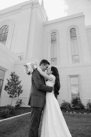 Black and white photo of a bride and groom embracing and kissing outside a grand white church building.