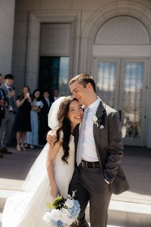 Smiling bride and groom embrace outside a grand building on their wedding day, bride holding blue hydrangea bouquet.