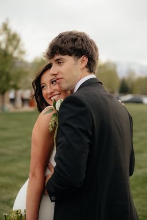 Bride and groom in intimate outdoor portrait, groom in black suit embracing bride in white dress on green lawn.