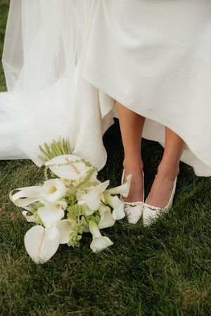 Bride wearing white heels stands beside elegant white calla lily wedding bouquet on green grass.