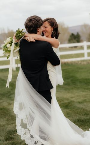 Groom lifts laughing bride in a white lace-trimmed veil near a white fence, as she holds a green bouquet.