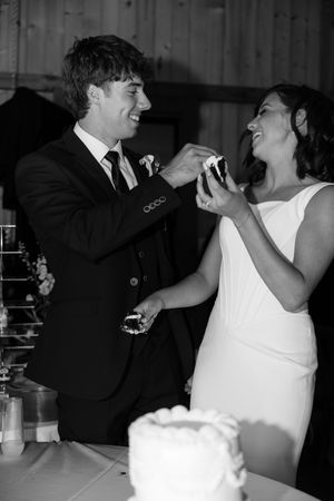 Bride and groom laugh while cutting their wedding cake at their reception in black and white photo.