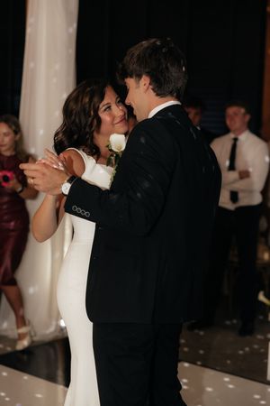Bride and groom share their first dance, she in a white gown, he in a black tuxedo with a white rose boutonniere.