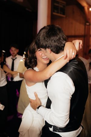 Bride laughs joyfully while embracing groom during wedding reception dance, wearing a sleeveless white gown.