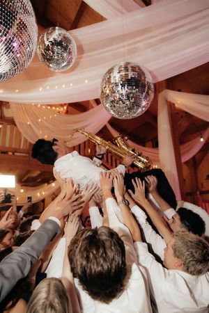 Excited wedding guests lift a saxophonist in white under glittering disco balls in a decorated barn reception venue.