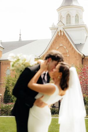 Bride and groom share a romantic kiss outside a historic brick church, with a white floral bouquet in the foreground.