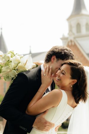 Groom in black suit kisses smiling bride holding white bouquet outside a church on their wedding day.