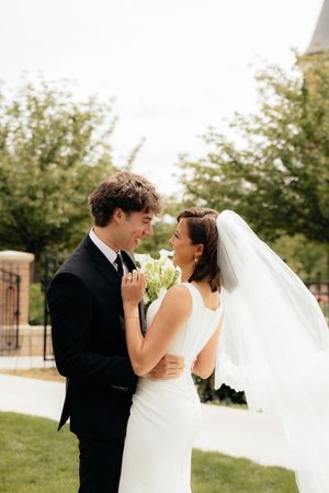 Bride and groom share a joyful moment outdoors, with the bride holding a white bouquet and veil flowing in the breeze.