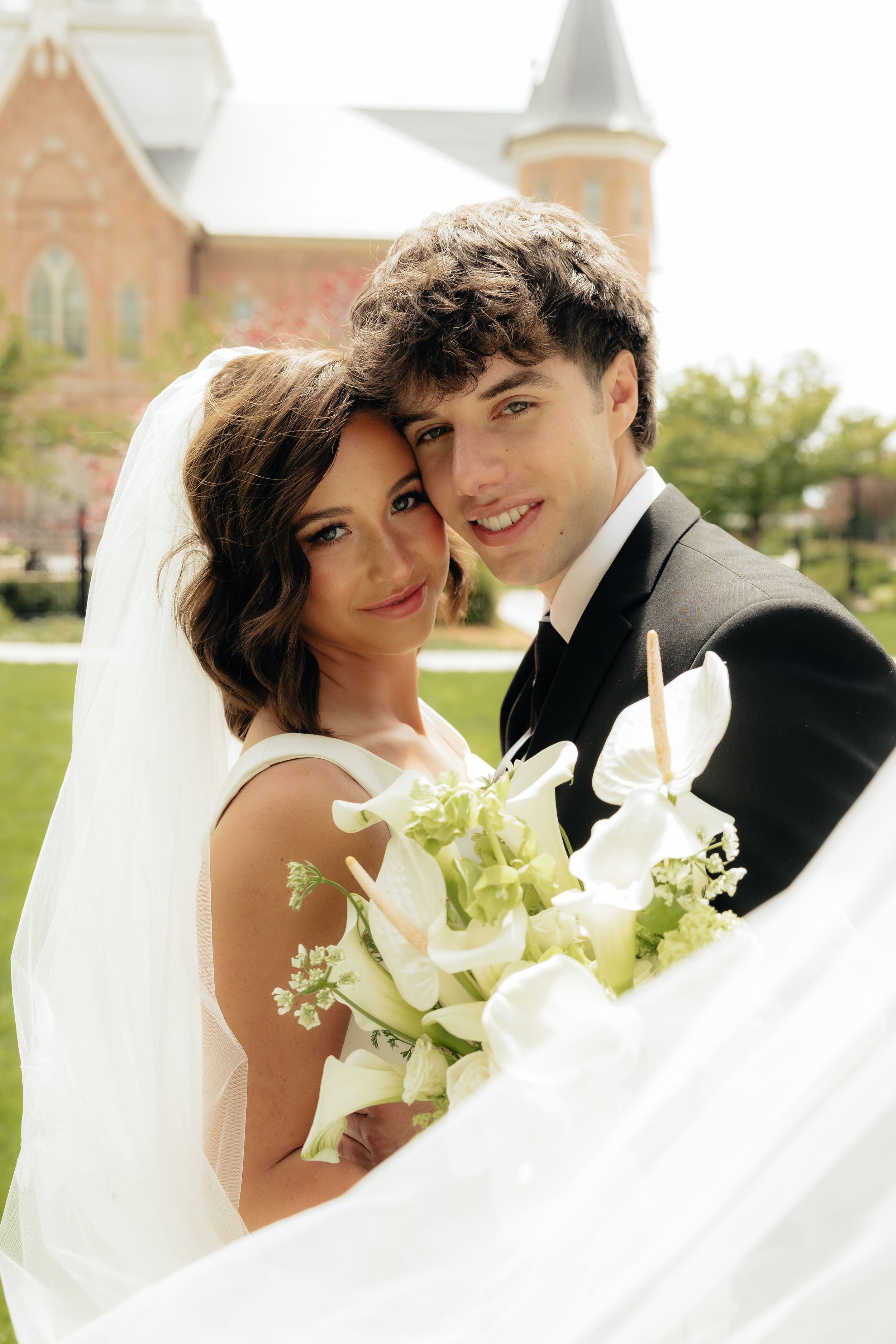 Bride and groom smiling together outdoors, bride holding white calla lily bouquet with veil flowing, church in background.