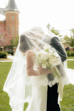 Bride and groom kissing under flowing lace-trimmed veil outdoors, bride holding white bouquet near historic church.