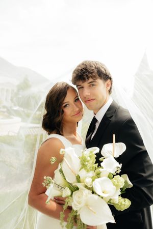 Bride and groom pose together under a veil holding a white floral bouquet on their wedding day.