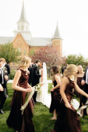 Blurred wedding party in brown bridesmaid dresses walks past a red brick church with white steeple on a cloudy day.