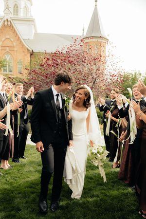 Bride and groom walk through petal toss with wedding guests cheering outside historic church.