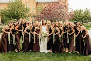 Bride poses with large bridal party in brown bridesmaid dresses on green lawn with red flowering tree backdrop.