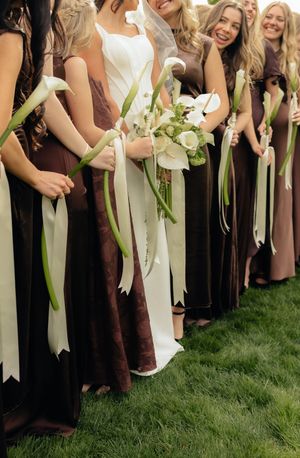 Bride stands with bridesmaids in brown dresses, all holding elegant white calla lily bouquets with long ribbon stems.