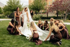 Bridesmaids in brown velvet dresses arrange bride's white wedding gown train on lush green lawn.