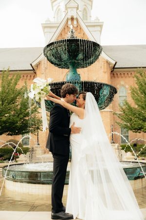 Bride and groom share a romantic kiss in front of a green fountain outside a brick church on their wedding day.