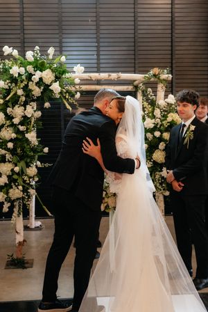 Bride and groom share first kiss at altar adorned with white floral arch during wedding ceremony.