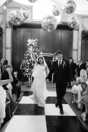 Bride and groom walk down checkered aisle celebrating after wedding ceremony under disco ball decorations.