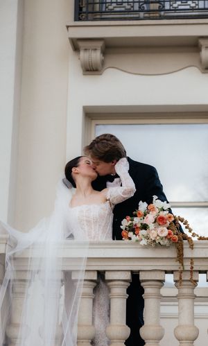A bride and groom share a romantic kiss on a balcony, surrounded by a beautiful architectural setting.