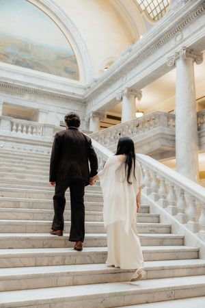 A couple, dressed formally, walk up the grand staircase of an ornate, historic building.
