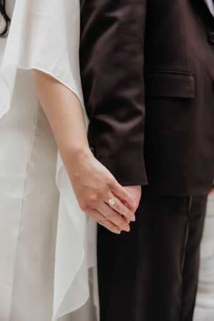 A close-up view of a bride and groom's hands clasped together, with the bride's white dress and the groom's dark suit visible.