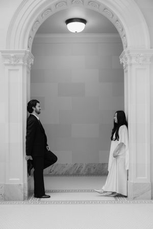 A man and woman stand facing each other in a formal, elegant interior with arched doorways and architectural details.