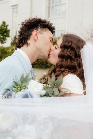 Bride and groom sharing a romantic first kiss, holding white and blue floral bouquet outdoors on their wedding day.