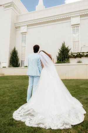 A bride and groom walk hand in hand toward a white temple, her long lace veil trailing on green grass.