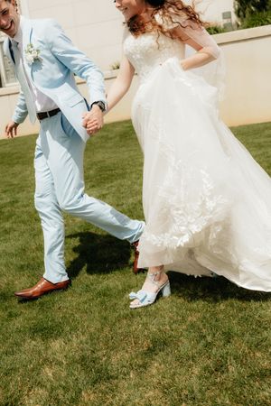 Bride in white lace gown and blue heels holds hands with groom in light blue suit running on green lawn.