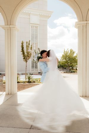 Bride and groom share a romantic embrace under white columns outside a grand temple on their wedding day.