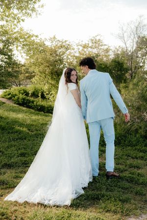 Bride in white gown with veil and groom in light blue suit walking hand in hand outdoors on green grass.