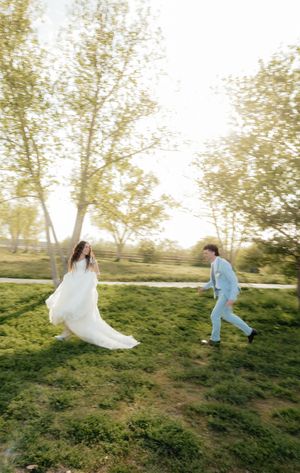 A bride in a flowing white gown and groom in a light blue suit joyfully run through a sunlit green park.
