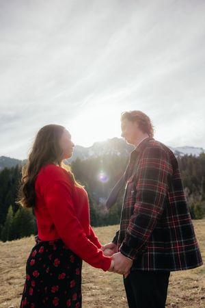 A couple holding hands and standing in a field with mountains in the background, with the sun shining brightly behind them.