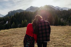 A couple stands together, facing a scenic mountain landscape with a red plaid blanket wrapped around them.