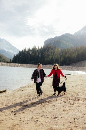Two people walking along a sandy beach with mountains in the background and a dog running beside them.
