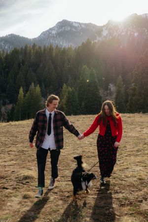 A couple walking hand-in-hand with their dog in a scenic mountain landscape with pine trees and sunlight.