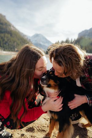 Two young women in red and black plaid shirts lovingly embrace a small black dog in a mountainous outdoor setting.