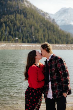 A young couple embracing and kissing in front of a scenic lake and forested mountains.