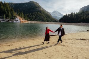 A couple walks hand-in-hand on a sandy beach near a serene lake surrounded by forested mountains.