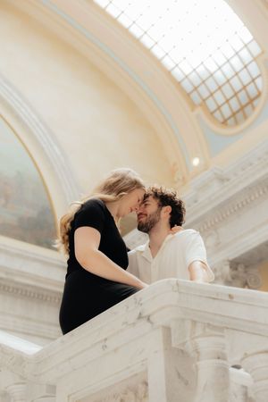 A couple embracing and kissing on a staircase in a grand, ornate building with arched windows.