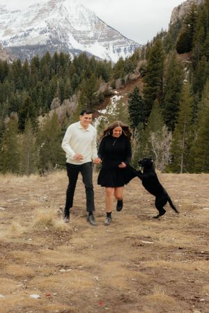 Two people walking together in a scenic mountain landscape with snowy peaks and pine trees.
