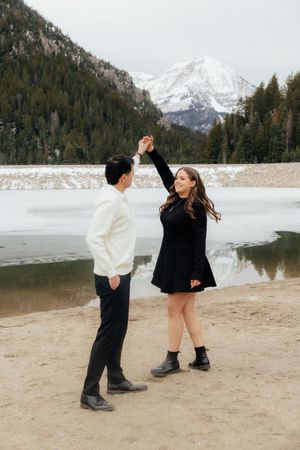 A couple stands on a beach near a snow-capped mountain and a frozen lake in a scenic winter landscape.