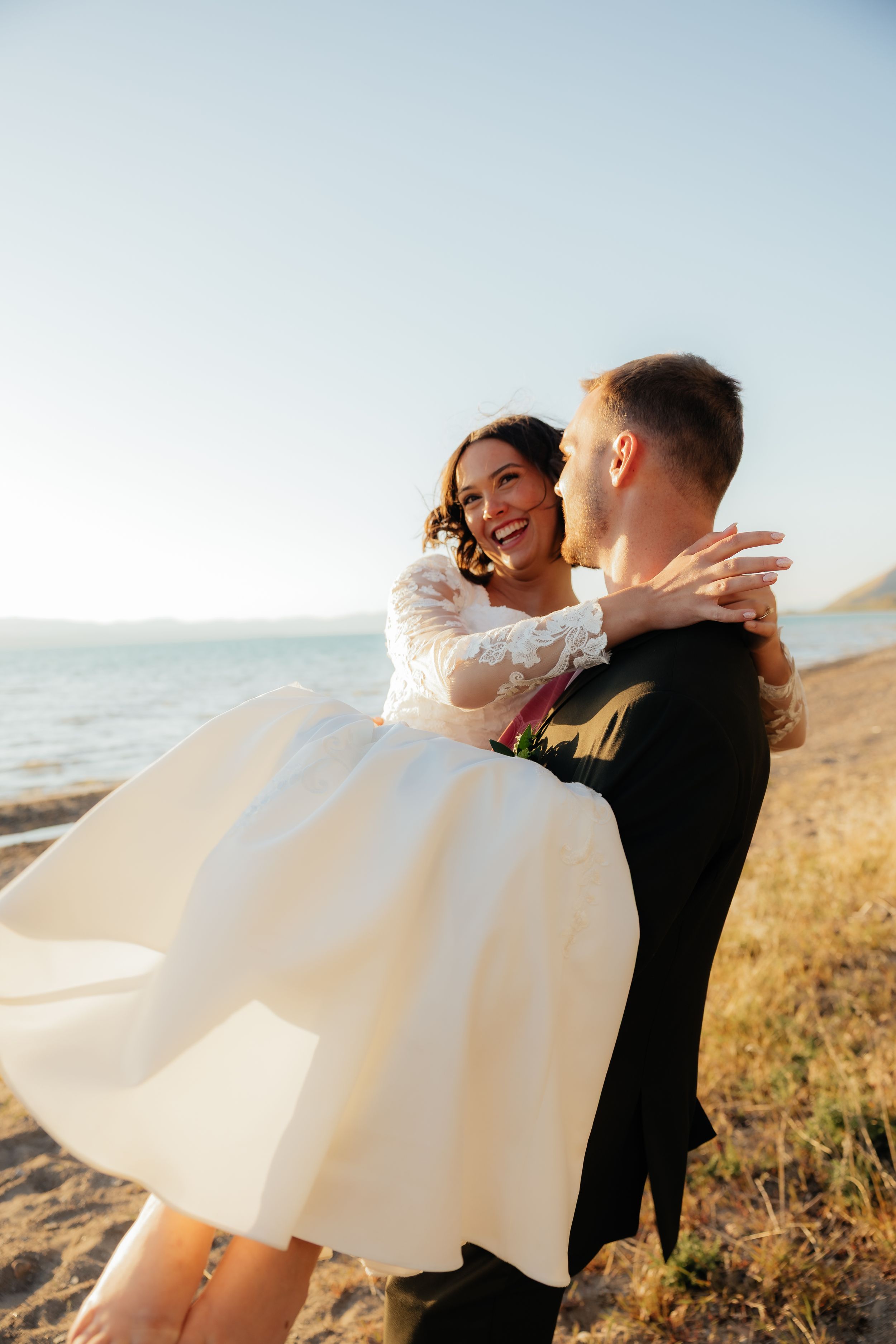 Groom lifting smiling bride in white dress on sandy beach with blue sky and water in background.