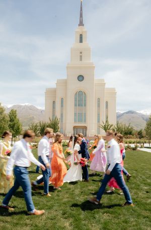 Wedding party walks past a white LDS temple with a spire, mountains visible in the background on a sunny day.