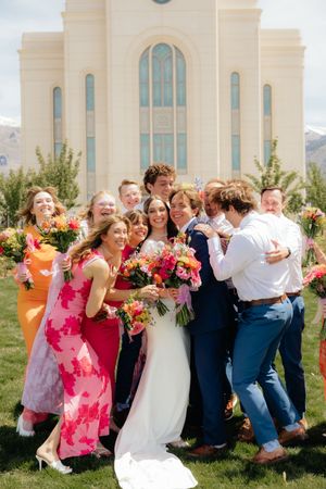 Bride and groom celebrate with wedding party in colorful dresses outside a white temple with mountain views.