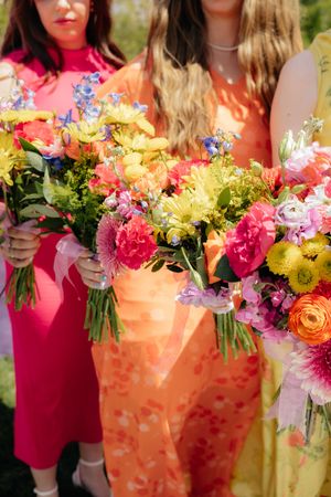 Three women in colorful dresses holding vibrant spring bouquets with pink, yellow, and orange flowers outdoors.