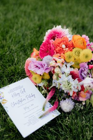 Colorful wedding bouquet with pink, orange, and purple flowers beside a wedding invitation on green grass.