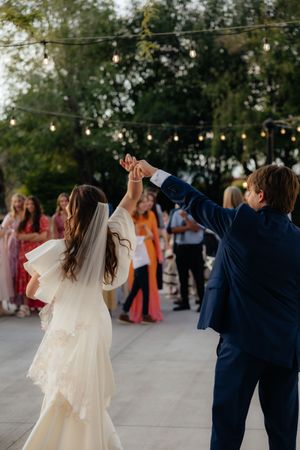 Bride and groom share first dance outdoors under string lights with wedding guests watching in background.