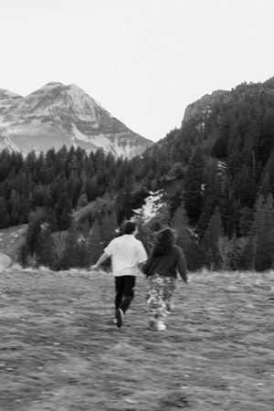 A black and white image showing two people walking through a shallow stream in a mountainous, forested landscape.